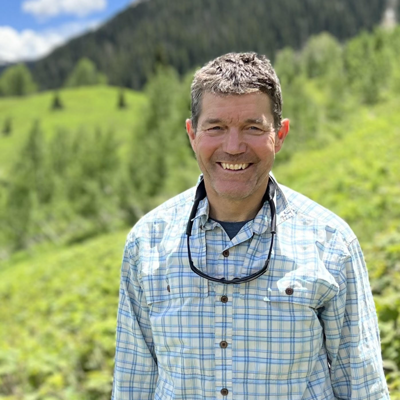 The image shows a smiling middle-aged man with short brown hair, wearing a blue and white checkered shirt and sunglasses around his neck. He is standing outdoors in a lush, green environment with trees and a hillside in the background. The lighting is bright and natural, suggesting a sunny day. The man appears relaxed and happy, enjoying the scenery.
